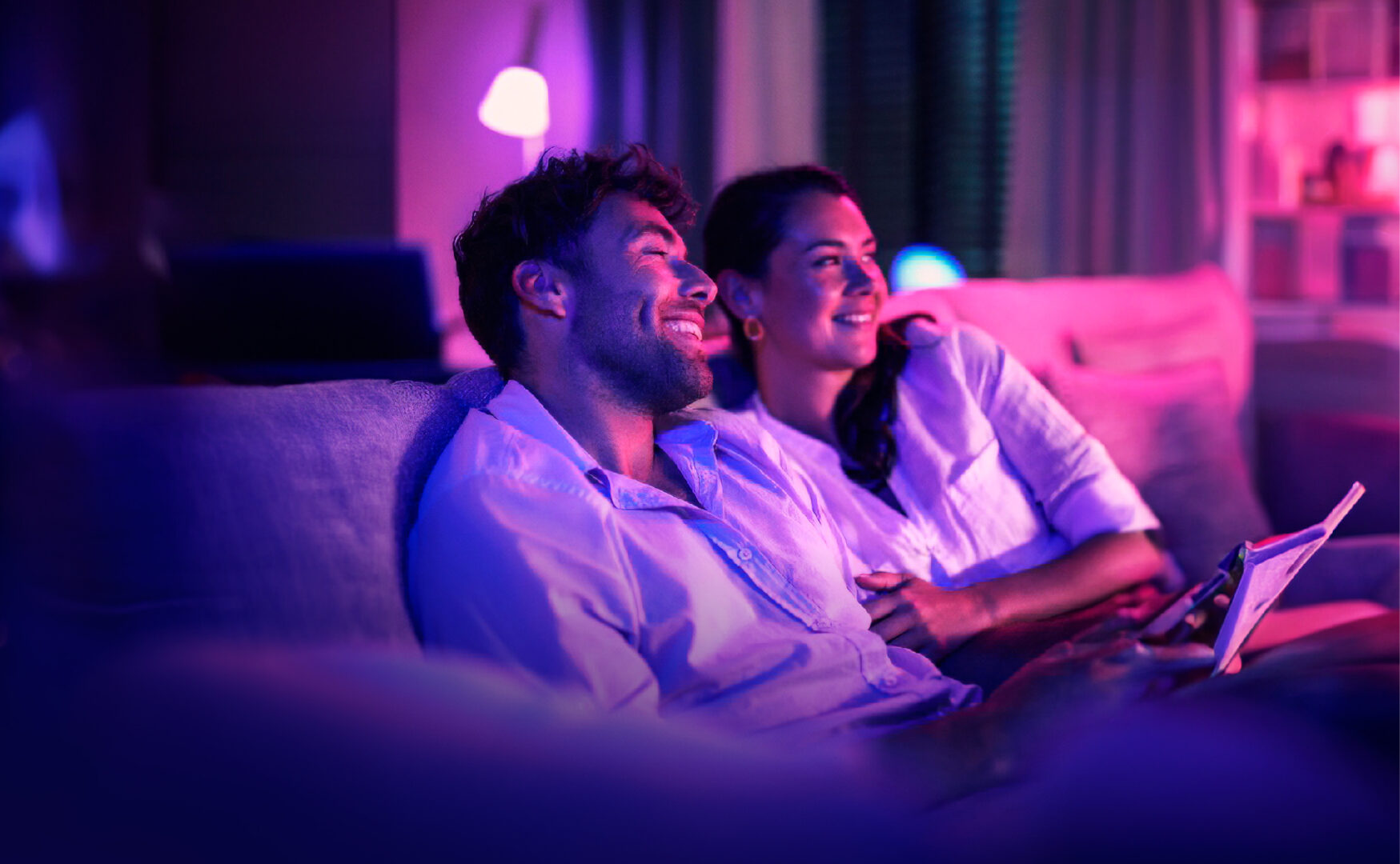 A couple sitting on the sofa watching the TV is a purple lit room