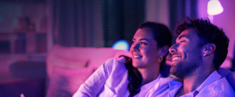 A couple sitting on the sofa watching the TV is a purple lit room